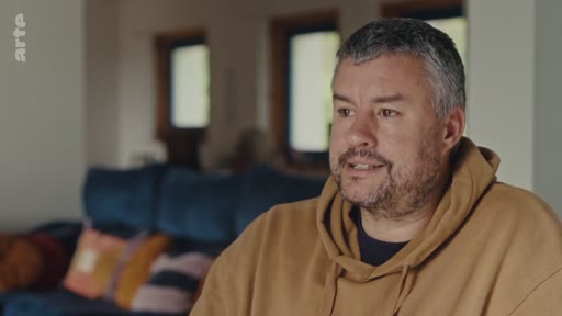 Un homme d'âge mûr, portant un sweat-shirt à capuche marron, est filmé en plan moyen lors d'une interview dans un intérieur domestique. Il s'exprime face caméra dans le cadre d'un reportage sur l'impact régional et l'expansion.