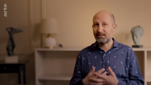 Un homme chauve avec une barbe s'exprime lors d'une interview sur le thème des campagnes de reforestation. Il est filmé en plan moyen dans un intérieur décoré avec des sculptures.