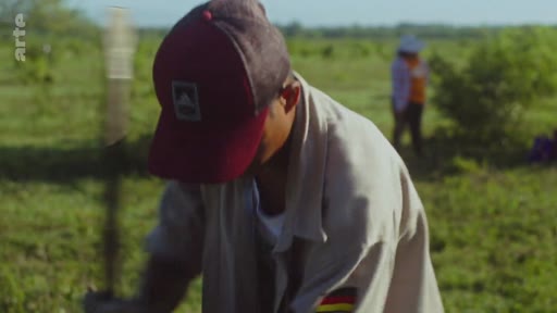 Un homme portant une casquette et une chemise avec un écusson travaille dans un champ en extérieur, illustrant les efforts de reforestation. En arrière-plan, une autre personne est visible dans ce paysage rural.