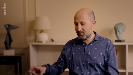 Un homme chauve avec une barbe, portant une chemise bleue à motifs, est interviewé dans une pièce décorée avec des sculptures et une lampe. Il participe à une émission sur le projet Plant for the Planet.
