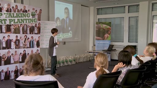 Un jeune garçon s'exprime devant un public lors d'une conférence organisée par l'initiative environnementale Plant-for-the-Planet. On peut voir une bannière avec le slogan 'Stop Talking Start Planting' et une projection montrant des visages de participants.