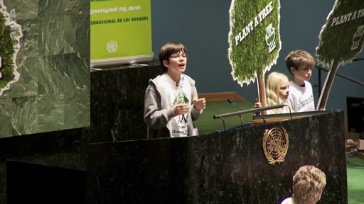 Un jeune garçon prend la parole à la tribune de l'Assemblée générale des Nations Unies, entouré d'autres enfants tenant des panneaux 'PLANT A TREE'. Cet événement souligne l'engagement de la jeunesse en faveur de la protection de l'environnement et de la reforestation.