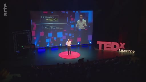 Un intervenant s'exprime sur scène lors d'une conférence TEDx à Lausanne, illustrant ses propos avec une infographie projetée sur grand écran mentionnant le 'total burden' de 300 Gt. La présentation aborde les enjeux environnementaux et le bilan carbone mondial.