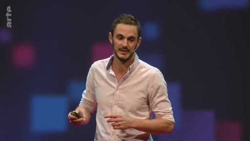 Un présentateur masculin s'exprime devant un public lors d'une conférence sur le climat, tenant une télécommande à la main. Il porte une chemise rose clair et se tient devant un fond sombre avec des motifs géométriques lumineux.
