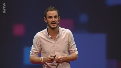 Un présentateur masculin s'exprime devant un public lors d'une conférence sur le climat, portant une chemise rose clair et un micro-cravate.