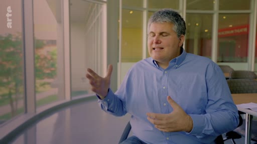 Un homme d'âge mûr, portant une chemise bleue, s'exprime avec des gestes de la main lors d'une interview filmée dans un espace de bureau moderne avec de grandes baies vitrées.
