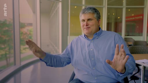 Un homme aux cheveux gris, vêtu d'une chemise bleue, gesticule en parlant lors d'une interview filmée dans un bureau moderne avec de grandes baies vitrées. Il participe à une analyse critique des méthodes scientifiques.