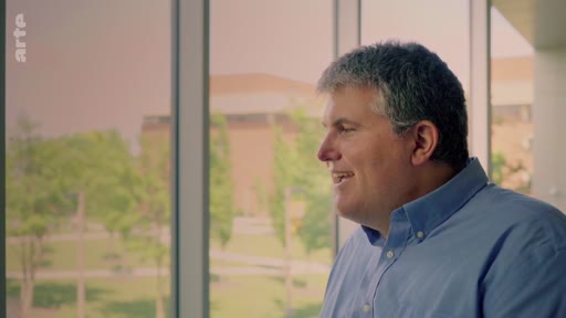 Un homme d'âge mûr, vêtu d'une chemise bleue, est filmé de profil lors d'une interview dans un cadre lumineux avec une vue sur un campus ou un parc en arrière-plan.