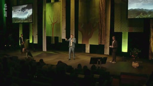 Un présentateur en costume s'exprime sur une scène de plateau télévisé décorée avec des panneaux illustrant des arbres et des paysages forestiers, lors d'une émission sur l'analyse critique des méthodes scientifiques.