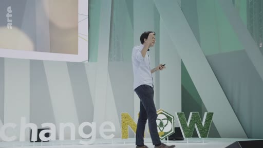 Un intervenant présente une conférence sur scène lors de l'événement ChangeNOW, devant un décor végétalisé. Il aborde les enjeux environnementaux et les implications géographiques de la plantation massive d'arbres.