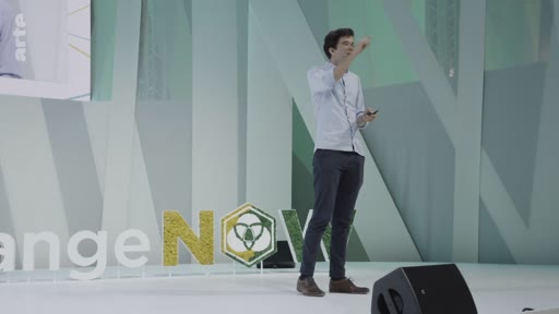 Un intervenant masculin présente une conférence sur la scène du sommet ChangeNOW, un événement international dédié aux solutions pour la planète. Il s'exprime devant un décor minimaliste portant le logo de l'événement.