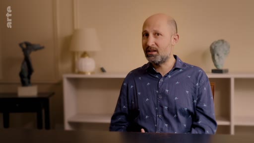 Un homme barbu, vêtu d'une chemise bleue à motifs, est interviewé dans un cadre intérieur sobre, évoquant les implications géographiques de la plantation massive d'arbres.