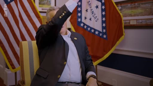 Un homme en costume s'exprime lors d'une interview, avec les drapeaux des États-Unis et de l'Arkansas en arrière-plan, dans le cadre d'un reportage sur le discours politique américain concernant le reboisement.