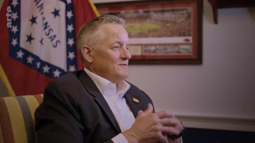 Un homme en costume est interviewé dans un bureau, avec le drapeau de l'État de l'Arkansas visible en arrière-plan, dans le cadre d'un reportage sur les défis de la neutralité carbone.