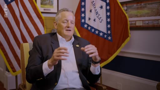 Un homme en costume, identifié comme un responsable politique, est interviewé devant les drapeaux officiels des États-Unis et de l'Arkansas. Il discute des enjeux liés à la neutralité carbone dans le cadre d'un reportage sur les défis environnementaux.