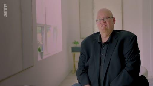 Un homme chauve portant des lunettes et une veste noire est interviewé dans un cadre intérieur lumineux, discutant probablement des enjeux liés à la compensation carbone.