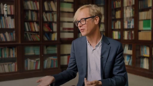 Un homme portant des lunettes et une veste de costume s'exprime lors d'une interview, avec une bibliothèque en arrière-plan, dans le cadre d'une discussion sur les critiques du marché du carbone et des pratiques des entreprises.