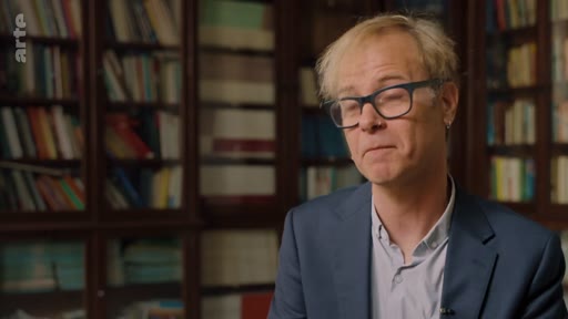 Un homme portant des lunettes est interviewé dans une bibliothèque, probablement pour un documentaire sur les enjeux environnementaux et les critiques des pratiques d'entreprise.