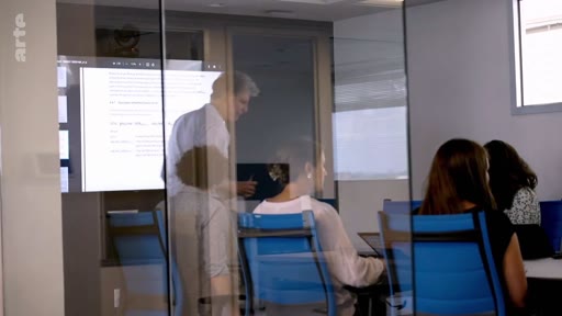 Un homme présente des documents projetés sur un écran lors d'une réunion de travail dans un bureau moderne avec des cloisons vitrées. Des collègues sont assis autour d'une table, écoutant la présentation dans un cadre professionnel.