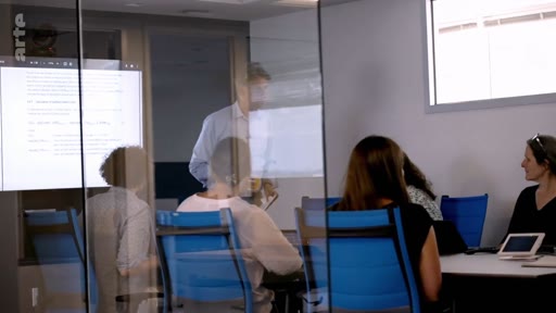 Une équipe de professionnels est réunie dans une salle de conférence vitrée pour une séance de travail, avec un écran de projection affichant des documents textuels en arrière-plan.