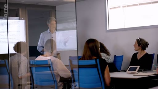 Un homme en chemise bleue fait une présentation devant un groupe de collaborateurs dans une salle de réunion moderne avec des parois vitrées. Le groupe discute de stratégies professionnelles ou de projets industriels.