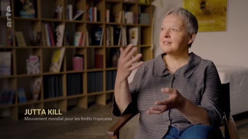 Jutta Kill, représentante du Mouvement mondial pour les forêts tropicales, est interviewée dans un cadre intérieur avec une bibliothèque en arrière-plan. Elle discute des enjeux liés à la protection des forêts tropicales et aux mécanismes de compensation carbone.