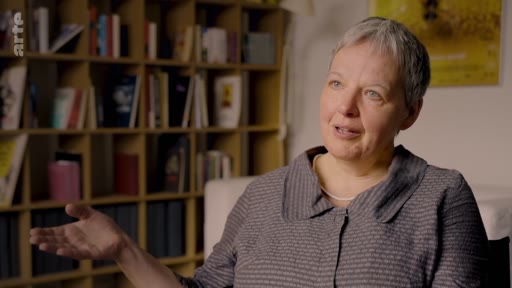 Une femme aux cheveux courts, interviewée dans un cadre intérieur devant une bibliothèque, s'exprime avec un geste de la main. L'image est issue d'un reportage sur la vérification et les incitations industrielles.