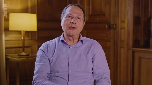 Un homme d'âge mûr, portant des lunettes et une chemise violette, est interviewé dans un cadre intérieur avec des boiseries et une lampe en arrière-plan, dans le cadre d'un reportage sur les pratiques industrielles.