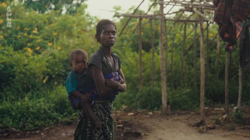 Une femme porte son jeune enfant sur le dos dans une zone rurale, illustrant la vie quotidienne et les défis des populations locales dans le cadre de la lutte pour les terres ancestrales.
