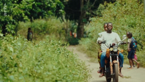 Un homme conduit une moto sur un chemin de terre dans une zone rurale, transportant des passagers, tandis qu'un enfant marche sur le côté. Cette scène illustre les conditions de transport et la vie quotidienne dans les zones rurales, souvent liées aux enjeux de développement et d'accès aux terres.