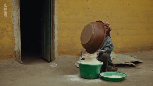 Une personne manipule des récipients contenant de la farine devant une maison aux murs jaunes, illustrant les conditions de vie et les activités quotidiennes dans une zone rurale.