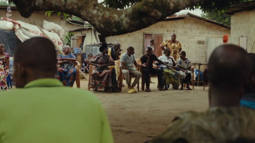 Des membres d'une communauté locale sont réunis en extérieur pour une discussion, assis sur des chaises sous un arbre, illustrant la vie sociale et les échanges au sein d'un village.