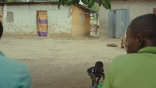 Un jeune enfant joue avec un objet dans la cour d'une habitation modeste en terre battue, sous le regard d'adultes au premier plan. Cette scène illustre les conditions de vie quotidiennes dans une zone rurale ou un quartier défavorisé.