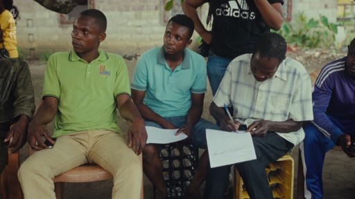 Un groupe d'hommes, dont un homme en chemise à carreaux qui prend des notes sur un document, est réuni en extérieur pour discuter de questions contractuelles ou administratives.