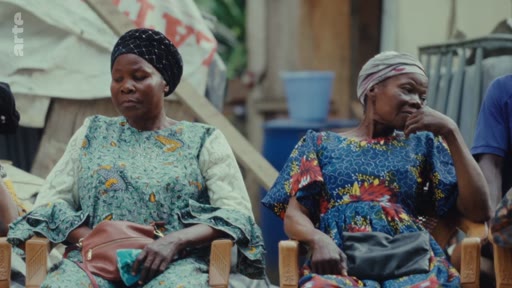 Deux femmes africaines vêtues de tenues traditionnelles sont assises côte à côte lors d'un rassemblement ou d'une réunion, illustrant des contextes de médiation ou de vie communautaire.