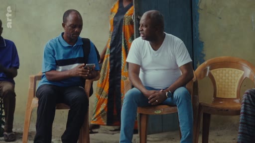 Deux hommes discutent assis sur des chaises en extérieur, l'un d'eux consultant son téléphone portable, dans le cadre d'un reportage sur des litiges contractuels et des manipulations.