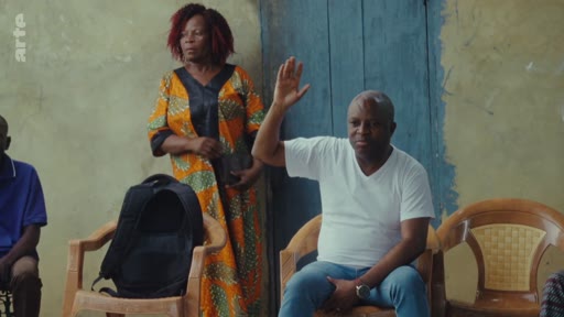 Un homme en t-shirt blanc lève la main pour prendre la parole lors d'une réunion, tandis qu'une femme en tenue traditionnelle se tient debout à ses côtés. La scène illustre une dynamique de groupe ou une assemblée dans un contexte de discussion ou de litige.