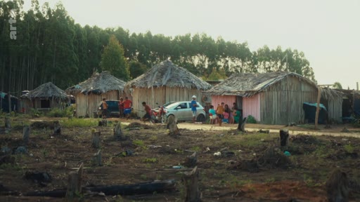 Vue d'ensemble d'un village composé de maisons en bois au toit de chaume, situé dans une zone forestière déboisée. Des habitants sont rassemblés autour d'une voiture garée devant les habitations, illustrant les conditions de vie dans une communauté indigène.