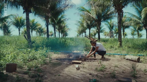 Une personne autochtone, portant une coiffe traditionnelle, cuisine en plein air sur un feu de camp dans une plantation de palmiers. Cette scène illustre les pratiques de subsistance et le mode de vie traditionnel au sein d'un environnement forestier.