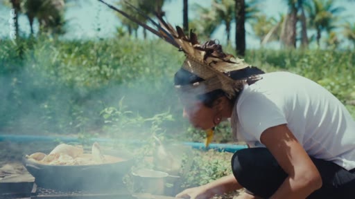 Une femme autochtone portant une coiffe traditionnelle prépare de la nourriture dans une marmite au-dessus d'un feu en extérieur, illustrant les pratiques culinaires et le mode de vie des peuples autochtones.