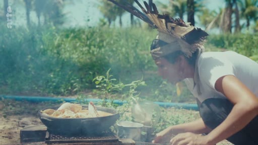 Une personne autochtone portant une coiffe traditionnelle cuisine dans une marmite posée sur un feu de camp en plein air, illustrant les pratiques culinaires et le mode de vie en forêt.