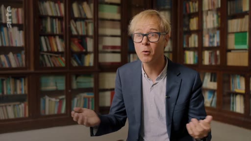 Un homme portant des lunettes et une veste de costume s'exprime lors d'une interview devant une bibliothèque remplie de livres, illustrant le thème de la recherche ou de l'expertise.