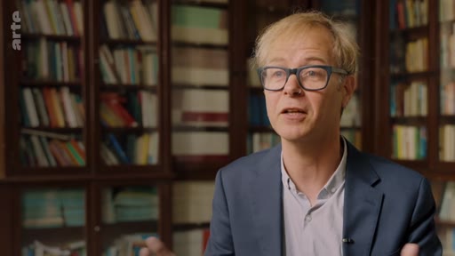 Un homme portant des lunettes et une veste de costume s'exprime face caméra lors d'une interview, avec une bibliothèque en arrière-plan. Il participe à un reportage sur la gestion forestière autochtone.