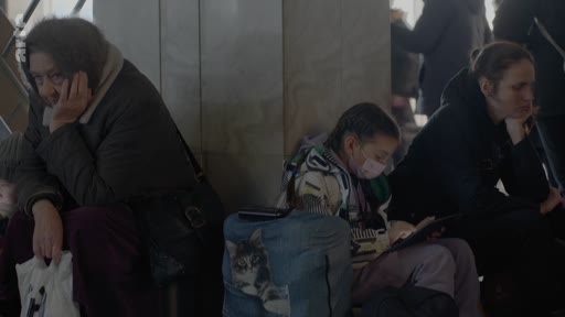 Des personnes, dont une femme âgée et une jeune fille portant un masque, sont assises dans une gare en attendant. Elles semblent fatiguées et portent des bagages, illustrant une situation de déplacement forcé ou de crise humanitaire.