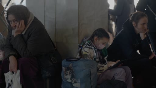 Des personnes, dont une femme âgée et une jeune fille, attendent dans une gare avec leurs bagages, dans un contexte de crise humanitaire ou de déplacement de population.