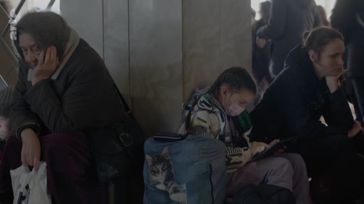 Des personnes, dont une femme âgée et une jeune fille avec une tablette, attendent dans une gare, illustrant une situation de crise humanitaire ou de déplacement de population.