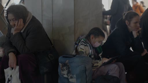 Des personnes, dont une femme âgée et une jeune fille portant un masque, attendent assises dans une gare. La scène évoque le contexte de l'exode ou du déplacement de populations civiles, possiblement en lien avec le conflit en Ukraine.