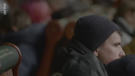 Un jeune homme portant un bonnet sombre est assis dans une salle d'attente, probablement dans une gare, entouré d'autres voyageurs. L'ambiance est calme et quotidienne, illustrant le transit des passagers.