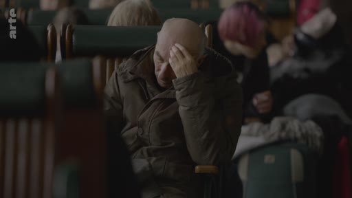 Un homme chauve d'âge mûr est assis dans un wagon de train, la main sur le front, semblant préoccupé ou épuisé. Il est entouré d'autres passagers dans un environnement de transport public.