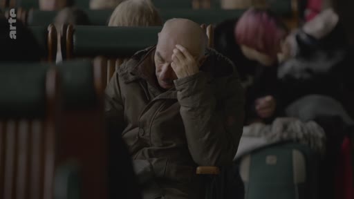 Un homme chauve d'âge mûr est assis dans un wagon de train, la main sur le front, semblant préoccupé ou fatigué. Il est entouré d'autres passagers dans une ambiance sombre et calme.